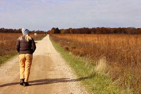Person on gravel path through tall grass field under cloudy sky